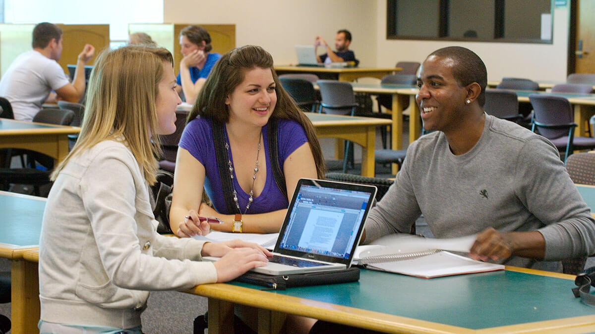 Three students at a table studying, one student is at a laptop, the other two are discussing notes.