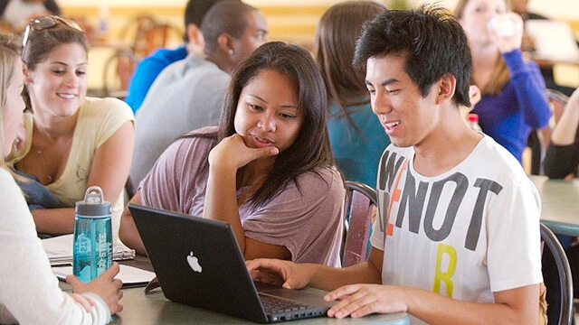 Two students sitting at a table with others, looking at a laptop computer